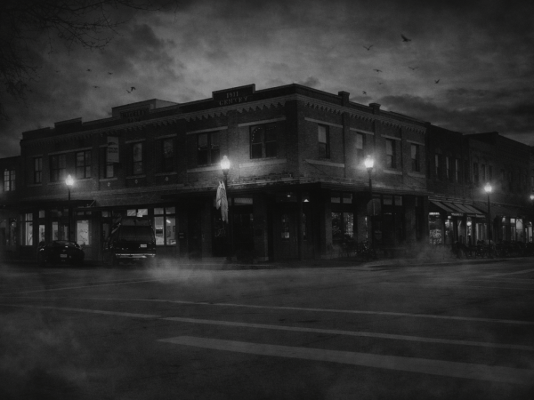 Black and white photo of a foggy street corner with historic buildings and streetlights at night.