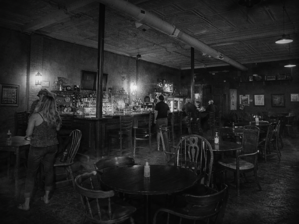 Black and white image of people in a dimly lit bar with wooden tables and chairs.