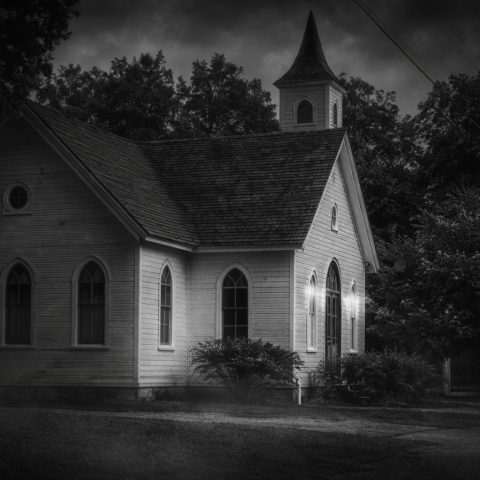 Old wooden church with steeple under cloudy, dark sky in grayscale.