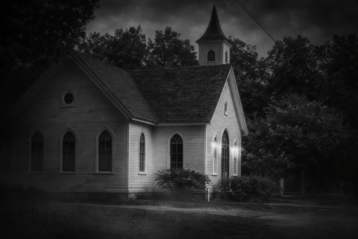 Old wooden church with steeple under cloudy, dark sky in grayscale.