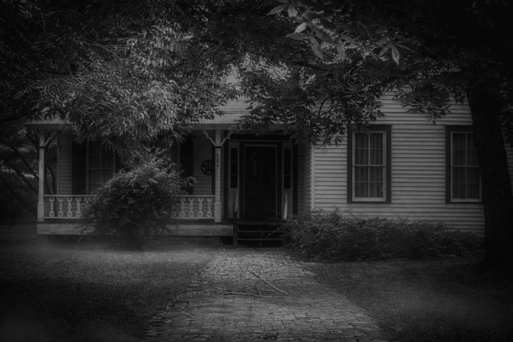 Foggy black and white image of an old house with a porch and overhanging trees.