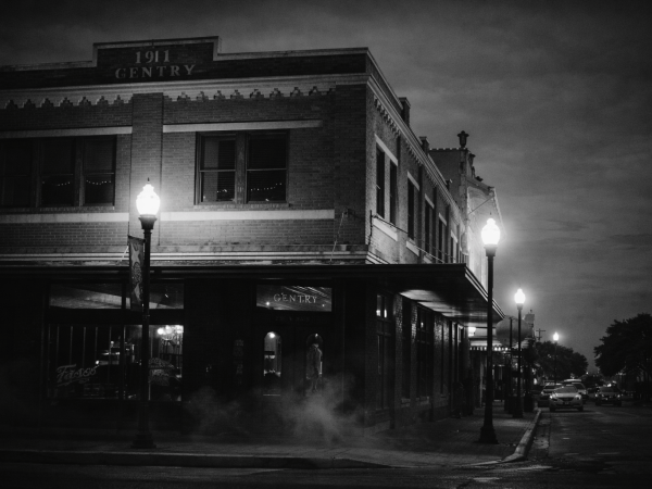 Black and white photo of a street corner with vintage buildings at night, lit by vintage-style street lamps.