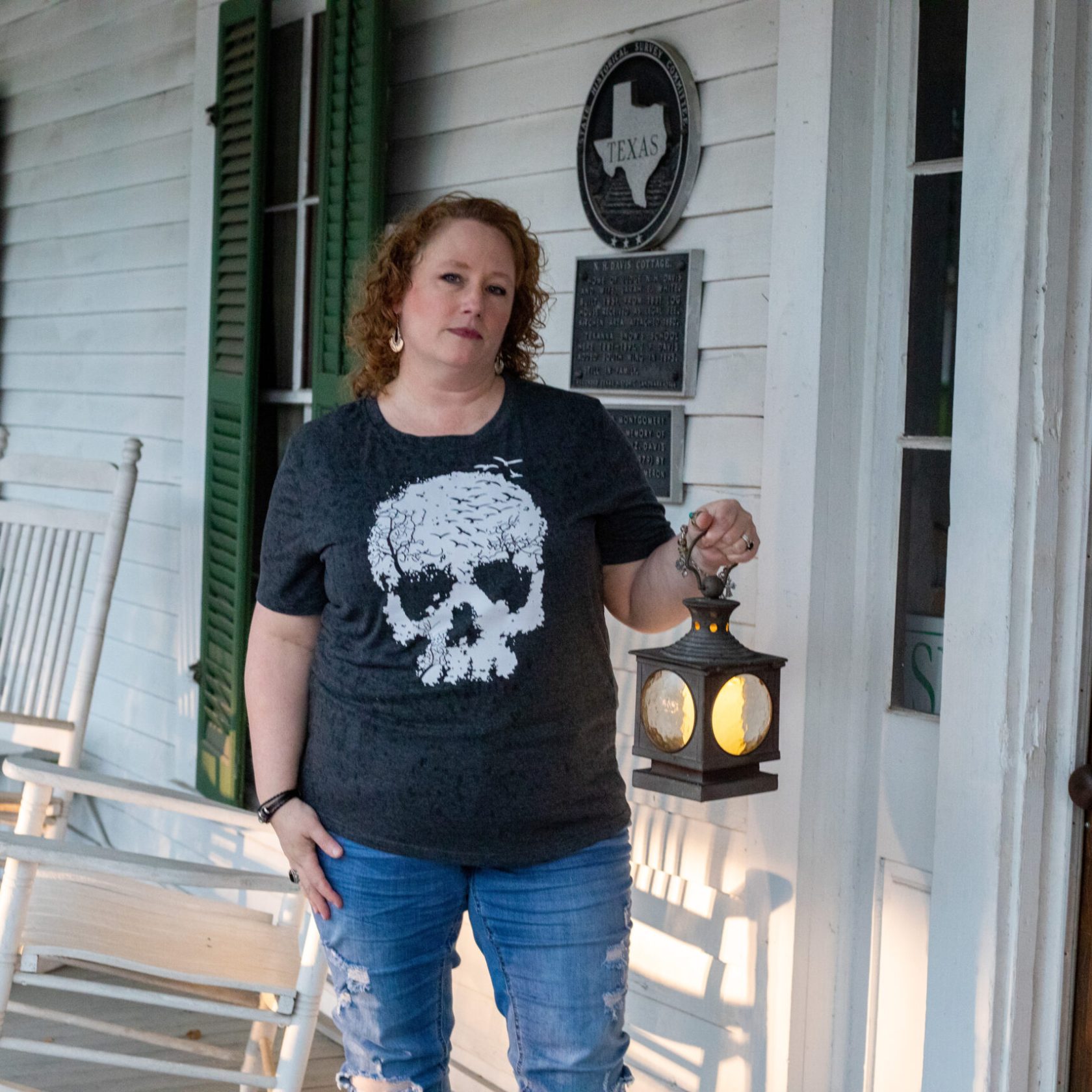 Person holding lantern on porch with rocking chairs and historical plaques.