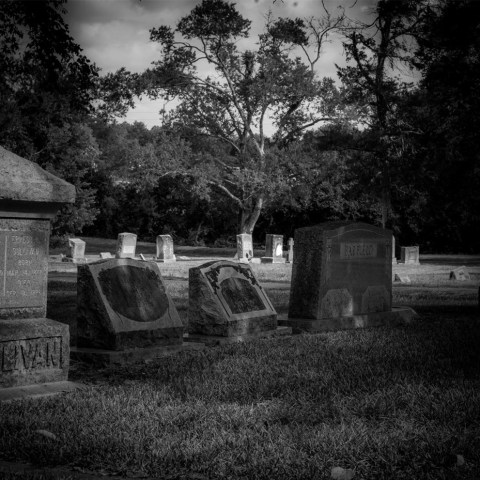 Black and white image of a graveyard with headstones, trees in the background.
