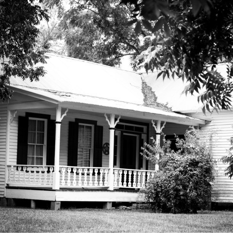 Black and white image of a vintage house with a front porch and trees.