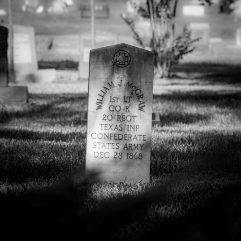 Black and white image of a gravestone in a cemetery, partially shadowed by surrounding trees.